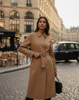 MiLA Boutique camel trench coat with cape detail, spread collar, belted waist, and buckle cuffs. Model stands on a cobblestone street.