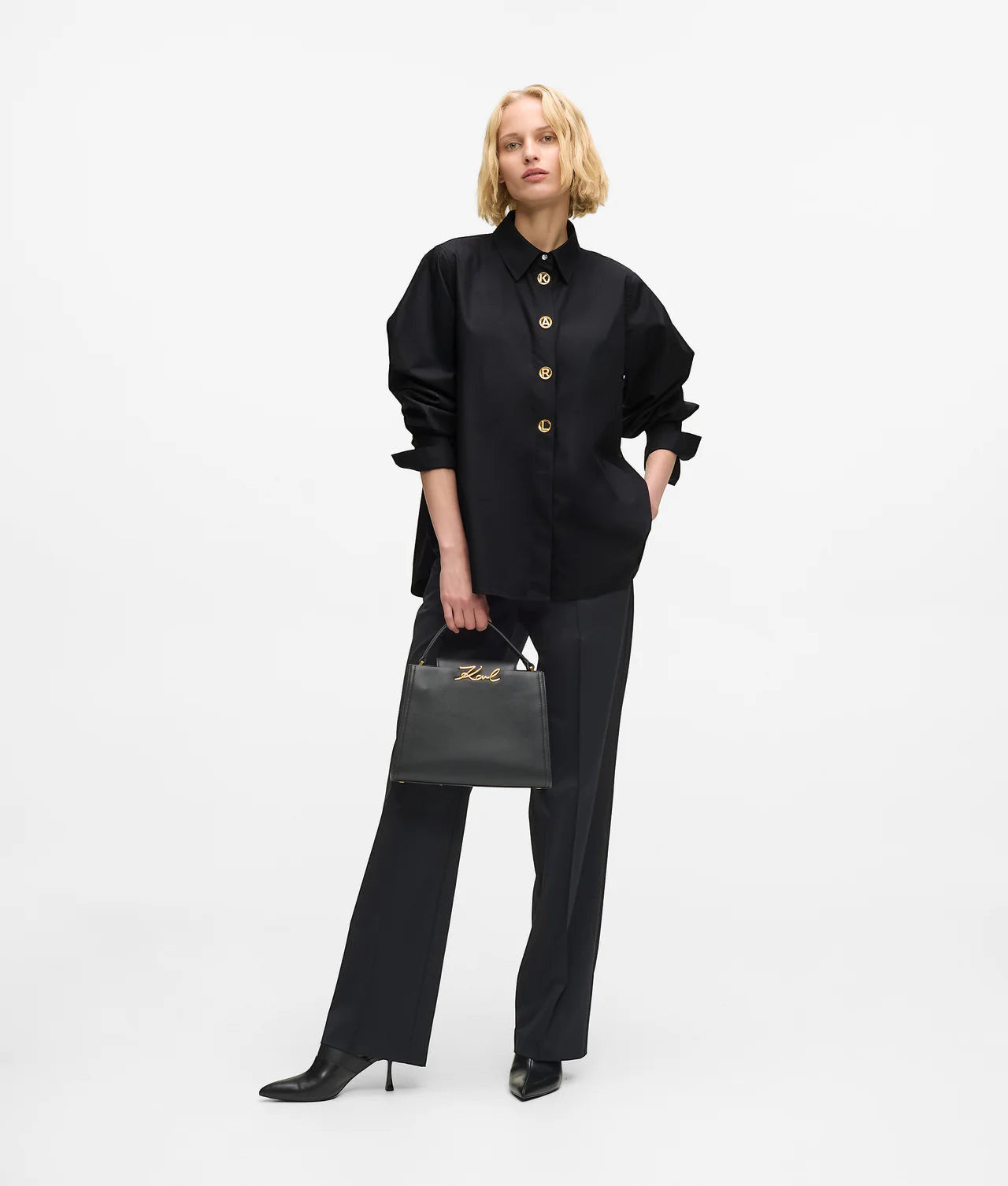 MiLA Boutique black cotton shirt with pointed collar and gold KARL lettering snap buttons, split side seams. Model holds a black KARL handbag.
