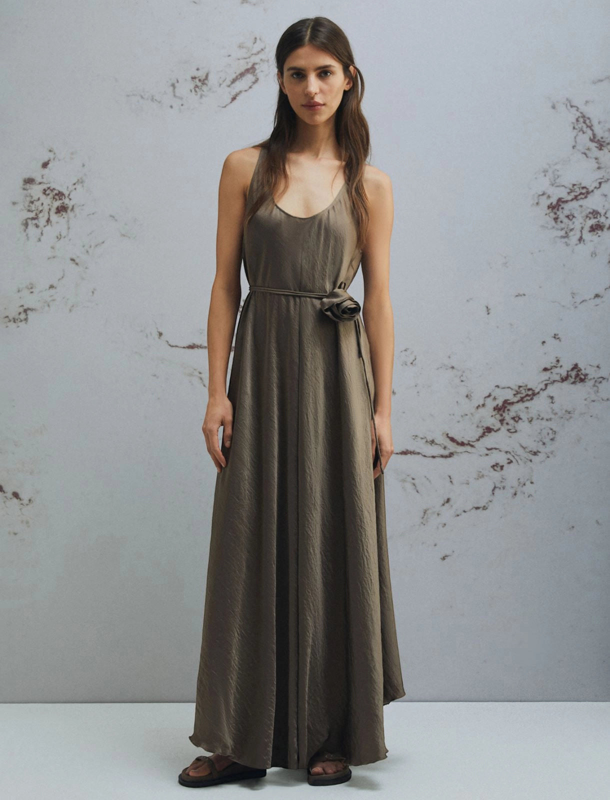 MiLA Boutique browncrinkle twill maxi dress with scoop neck, fabric belt, and flower detail. Model wears a flowing, asymmetrical hem dress.